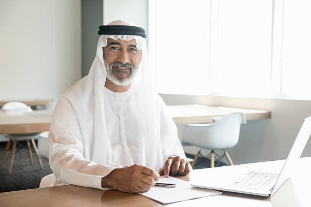 A photo of confident and smiling Emirati businessman with laptop. Portrait of professional is in traditional thobe. He is sitting at conference table, in a brightly lit office. Desk is found near windows. Dubai, United Arab Emirates, Middle East